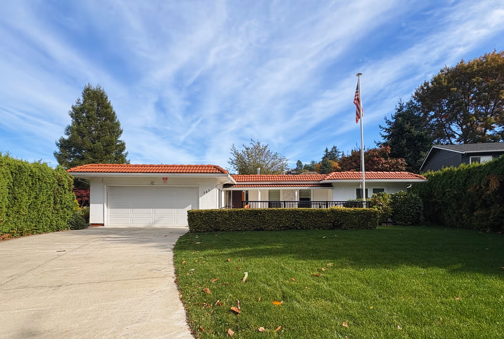 A single-story white house with a red-tiled roof, driveway, and flagpole in a green yard.