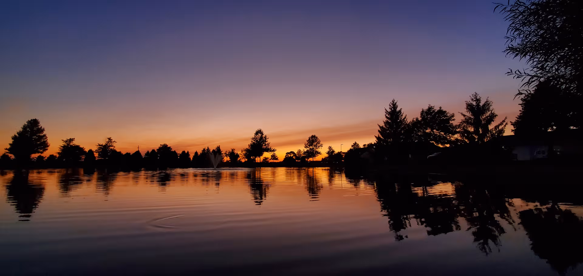 Sunset over a calm lake with silhouettes of trees and distant houses.