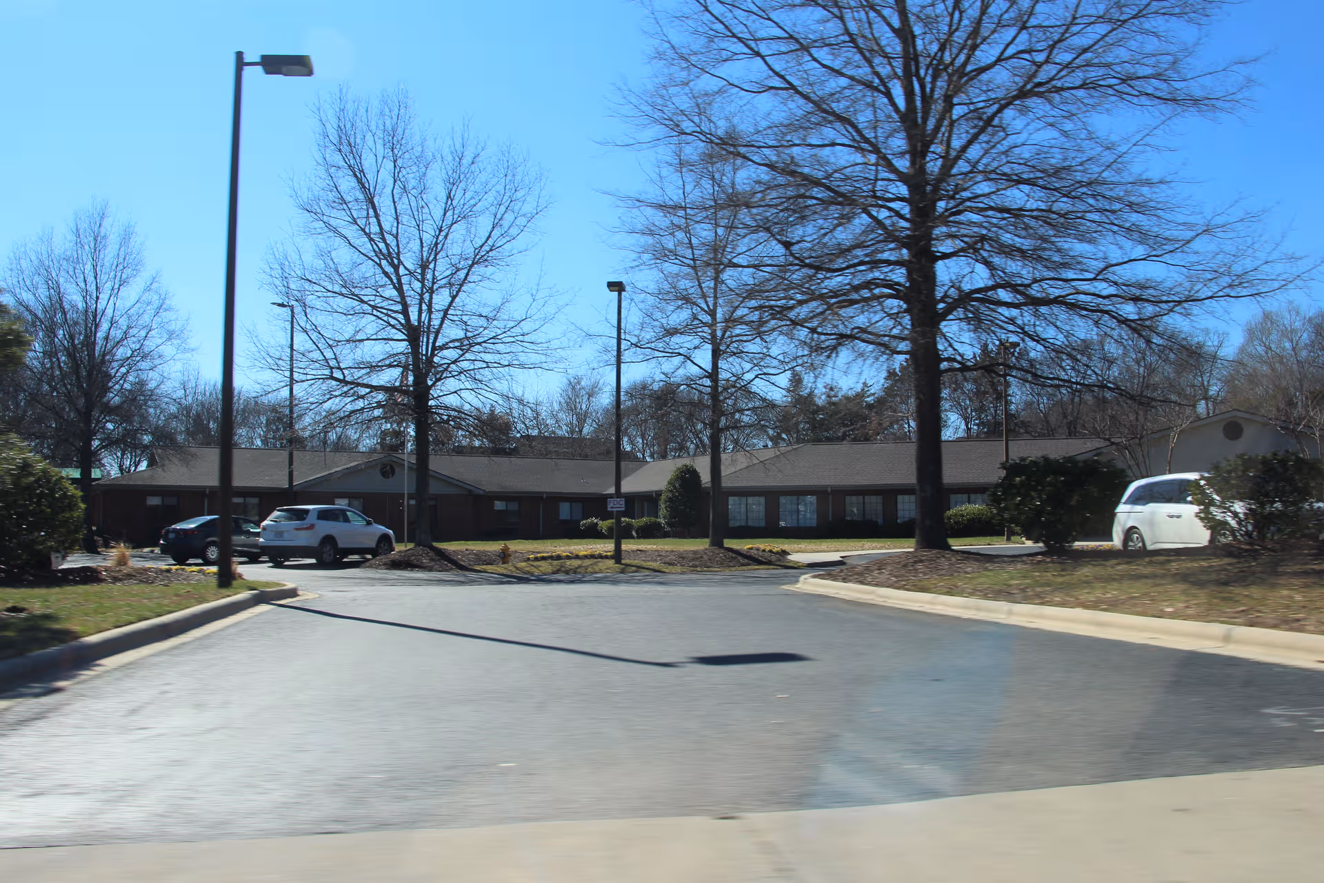A single-story brick building front with a parking lot and bare trees under blue sky.