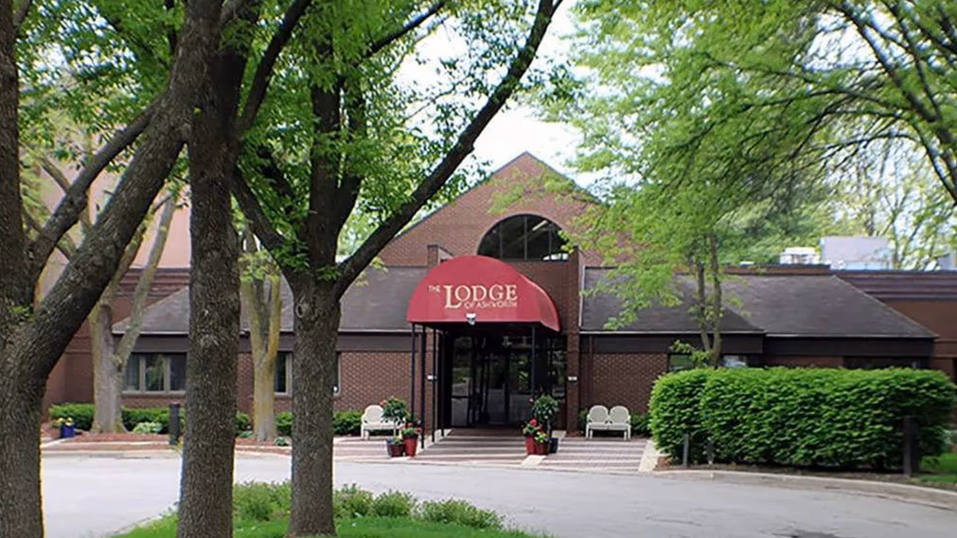 Brick building front with a red awning reading 'The Lodge' over the entrance, framed by trees, benches, and shrubbery.
