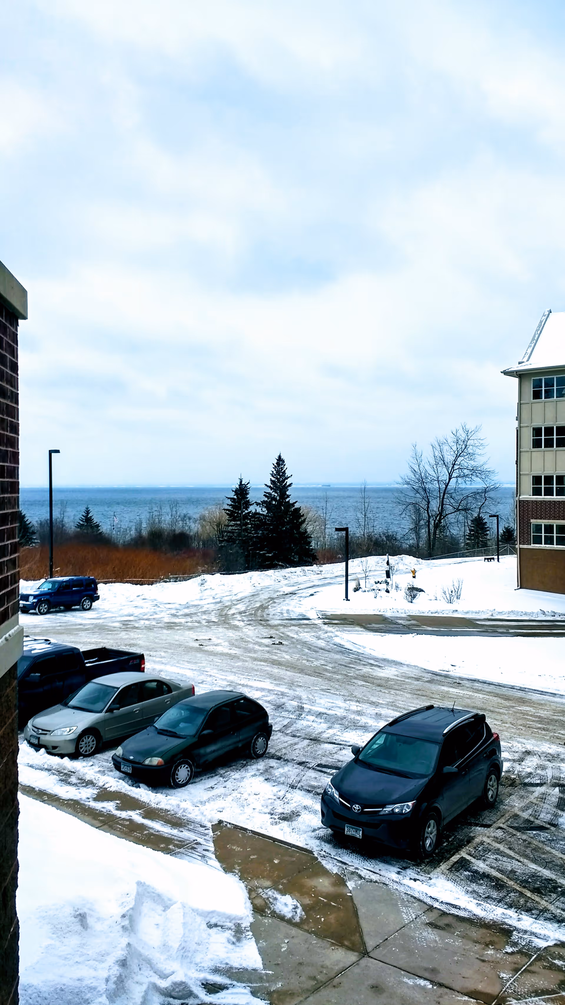 Snowy outdoor parking area with cars and distant water view.