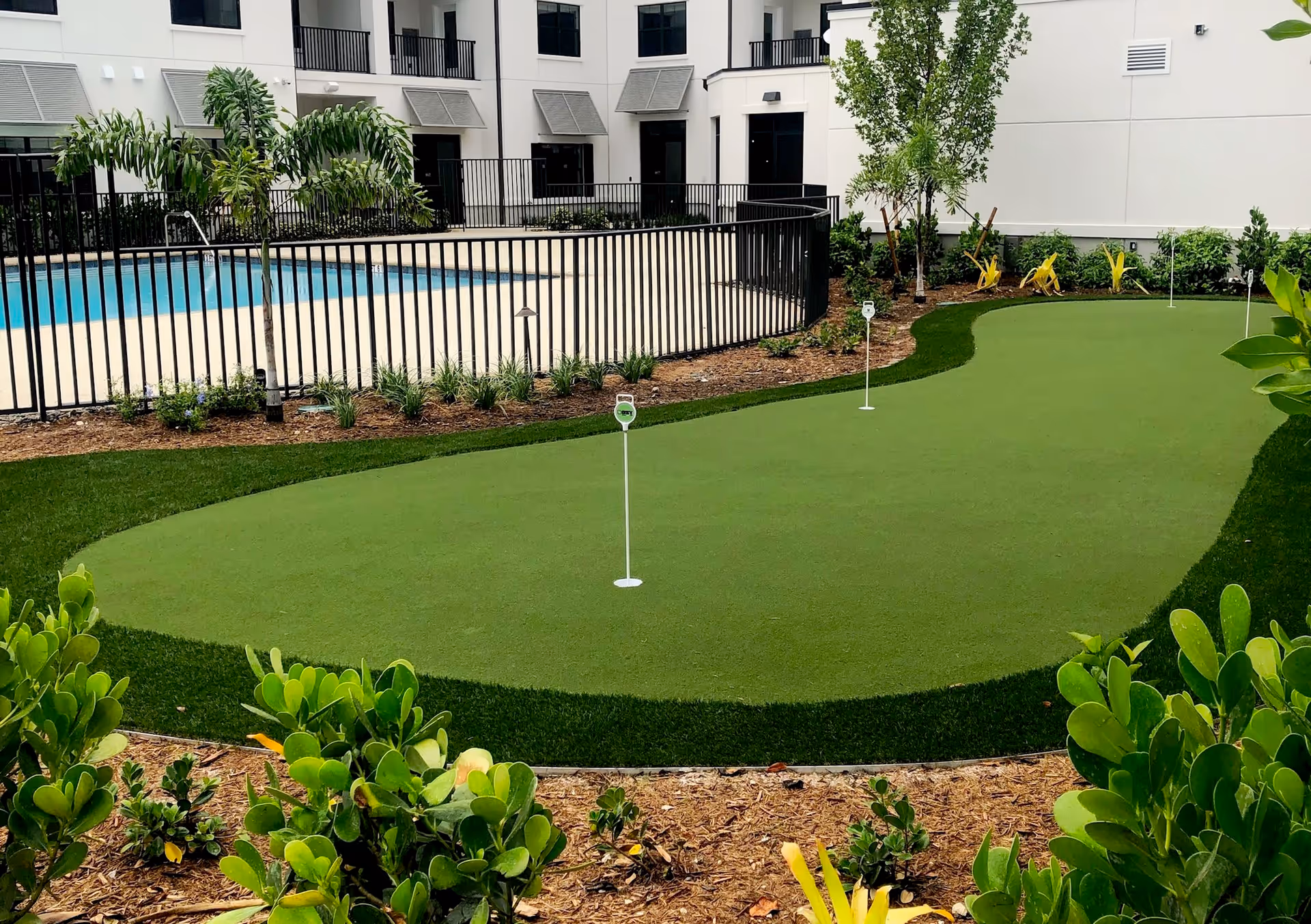 Outdoor area at Seascape At Naples featuring a putting green with three golf holes and flags, surrounded by landscaping with bushes and small trees. In the background, there is a fenced swimming pool adjacent to a white building with balconies and windows.