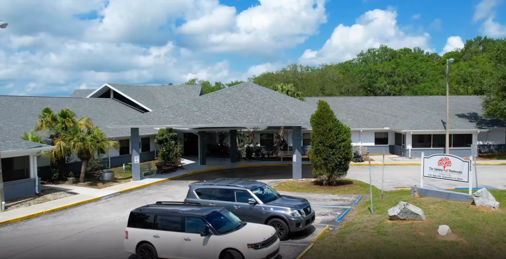 Exterior view of Gardens of Westbrooke senior living facility showing a single-story building with a covered entrance, surrounded by greenery and parked cars in front under a partly cloudy sky.