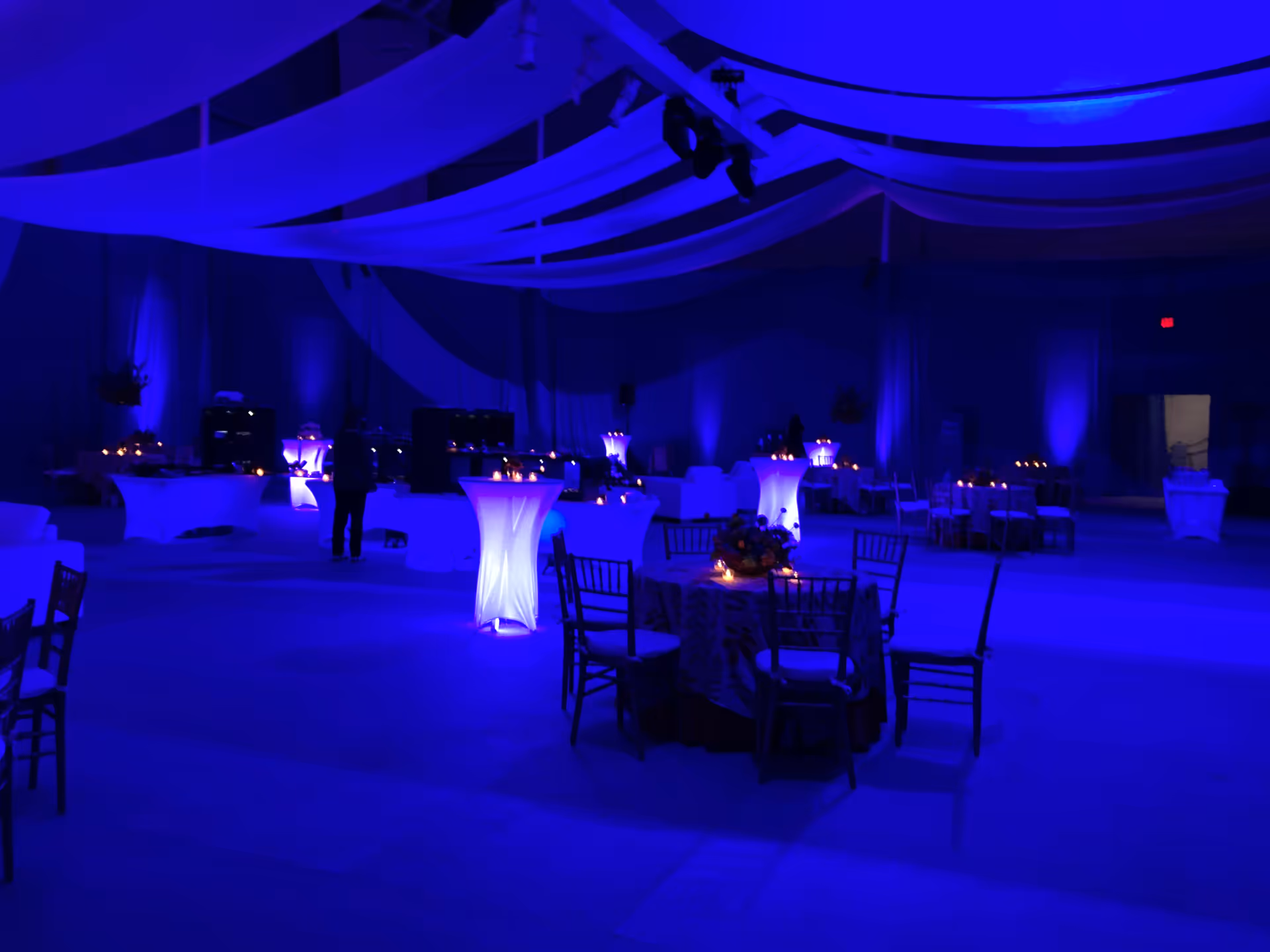 Interior banquet hall with blue uplighting, draped ceiling fabrics, and candlelit round tables.