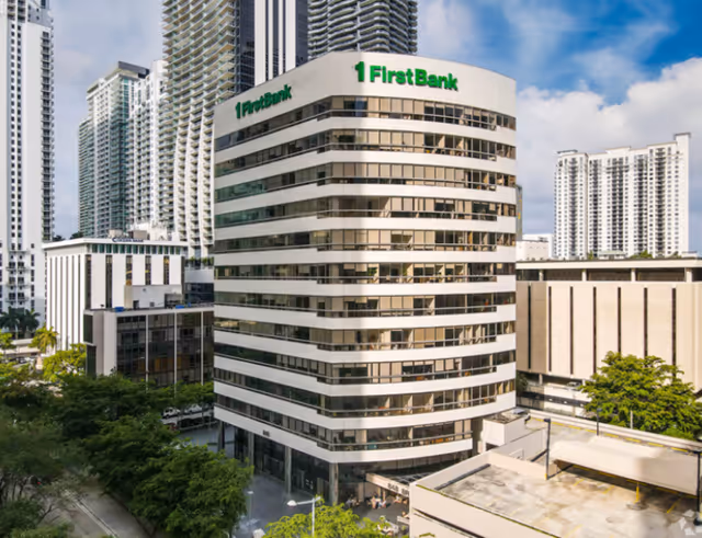 Curved white FirstBank building with green signage among tall surrounding skyscrapers.