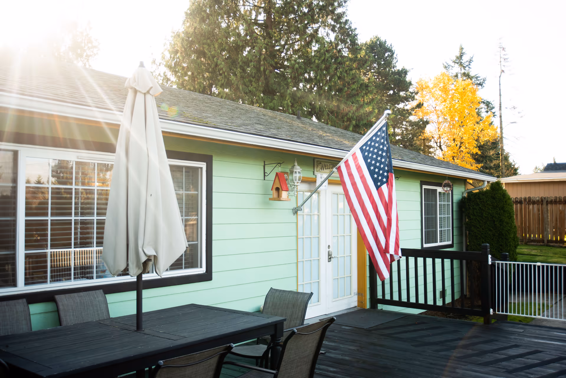 Mint-green house exterior with a wooden deck, outdoor dining table, umbrella, and American flag.
