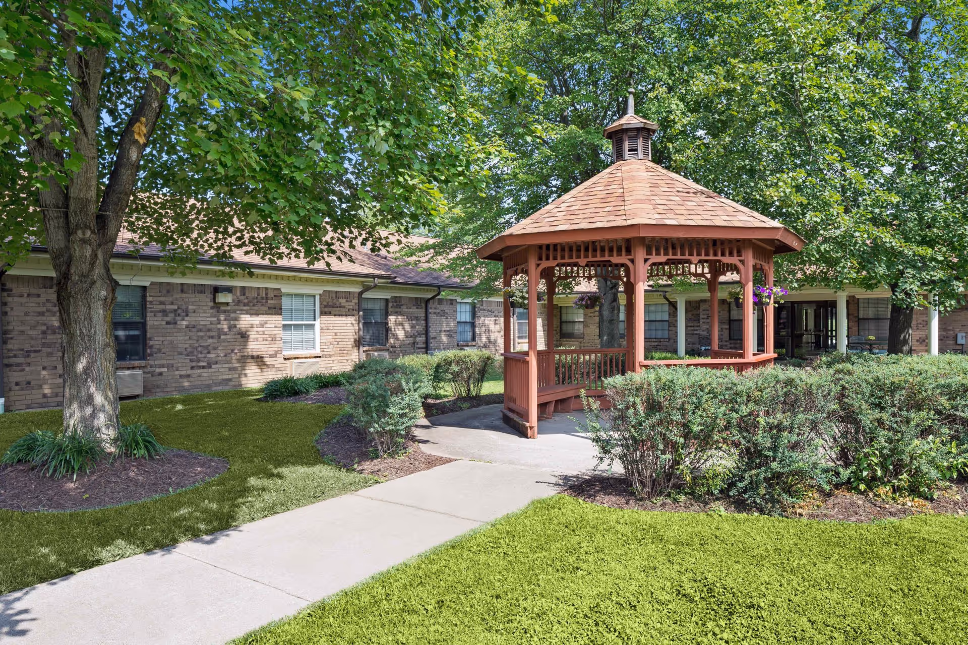 Outdoor view of a senior living facility showing a wooden gazebo surrounded by green bushes and trees, with a brick building in the background and a concrete walkway leading to the gazebo.