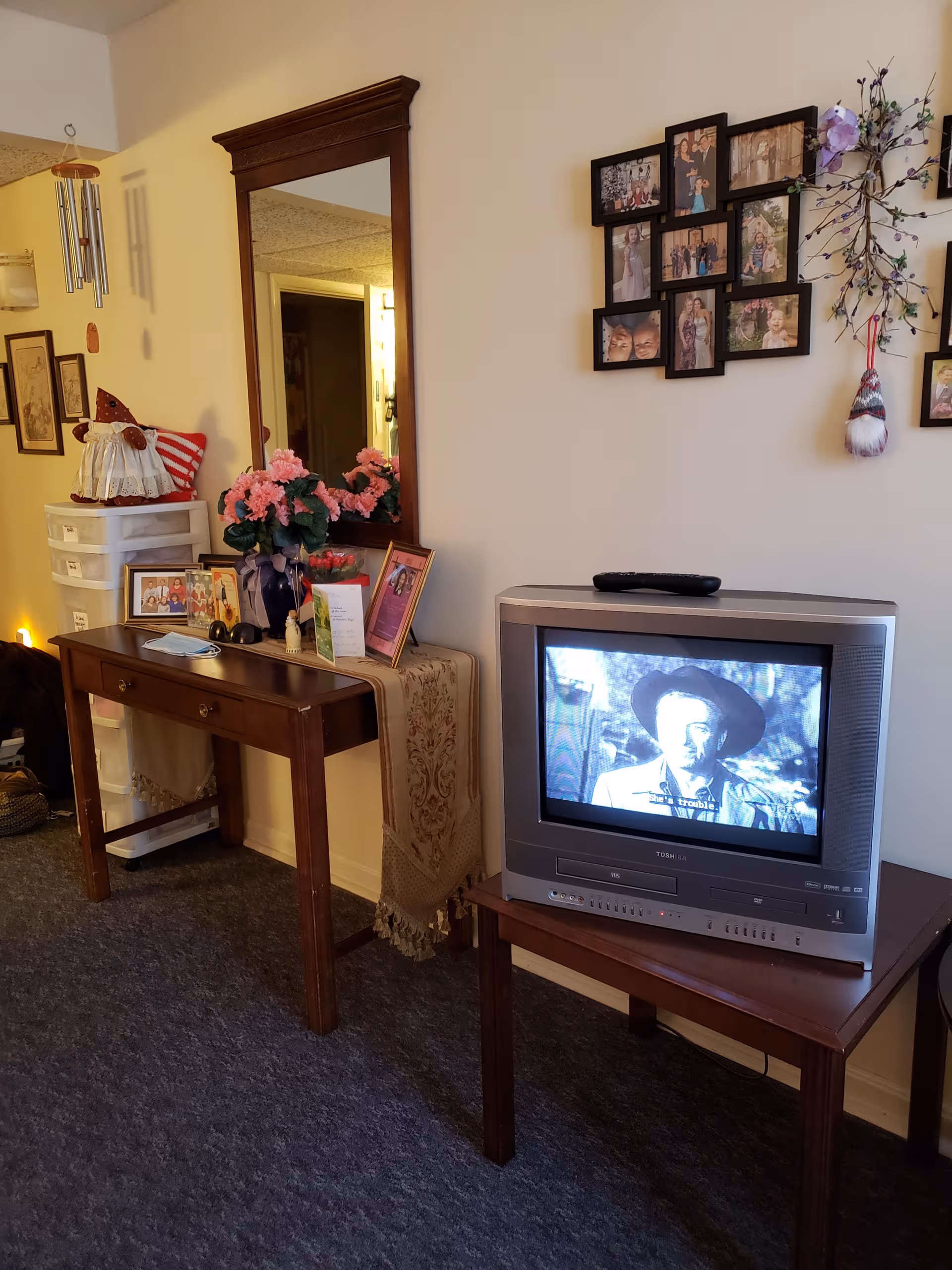 A cozy living area with a TV, mirror, photos, flowers, and a small dresser.