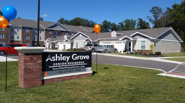 Exterior view of Ashley Grove Senior Residence showing a brick sign with the facility name and contact information in front of a driveway and several single-story residential buildings with parked cars and balloons.
