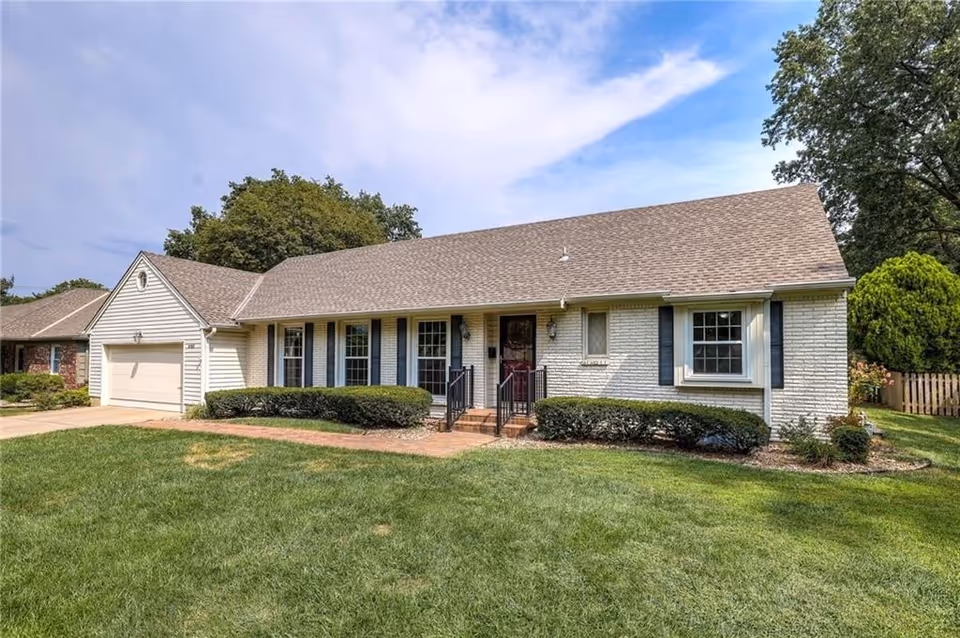A single-story white brick house with a two-car garage, a small front porch with steps, and a well-kept lawn.