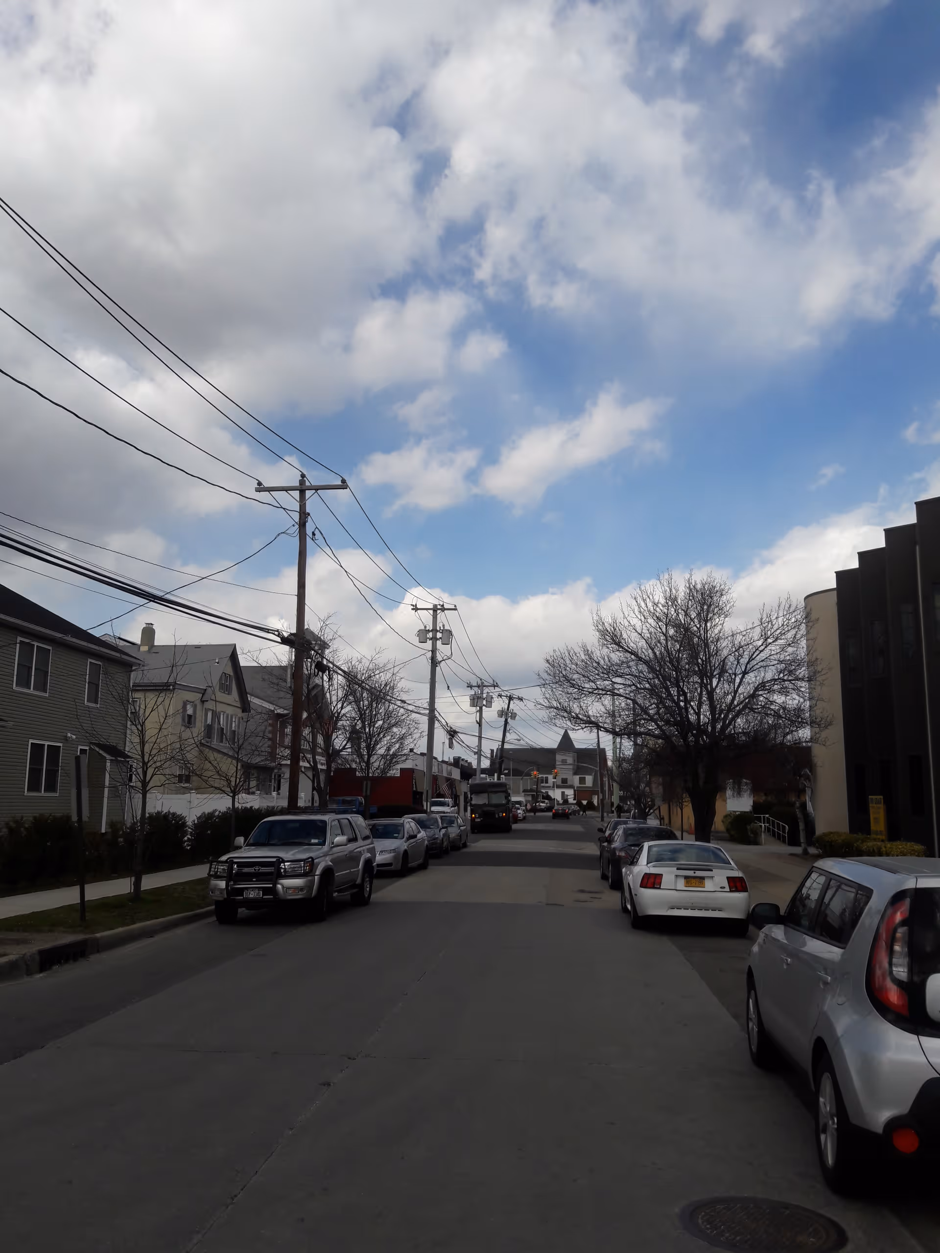 Residential street with parked cars, utility poles, and bare trees under a partly cloudy sky.