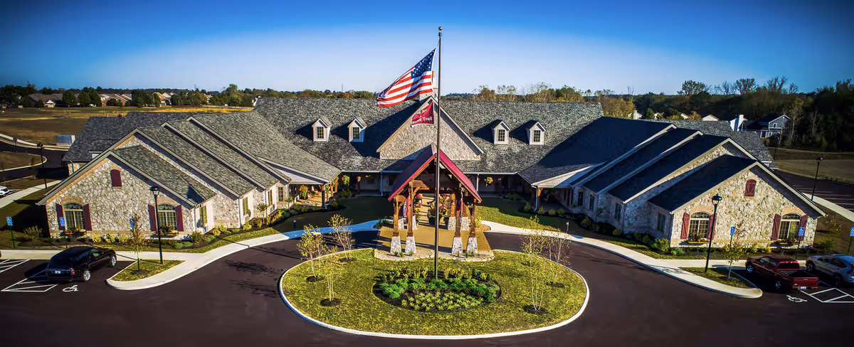 Aerial view of The Inn at Walnut Trail, a large stone building with a gray shingled roof and multiple dormer windows. In front of the building is a circular driveway with a landscaped island featuring a flagpole flying the American flag and a facility flag. Several cars are parked in the parking lot surrounding the building, and the area is surrounded by trees and open land under a clear blue sky.