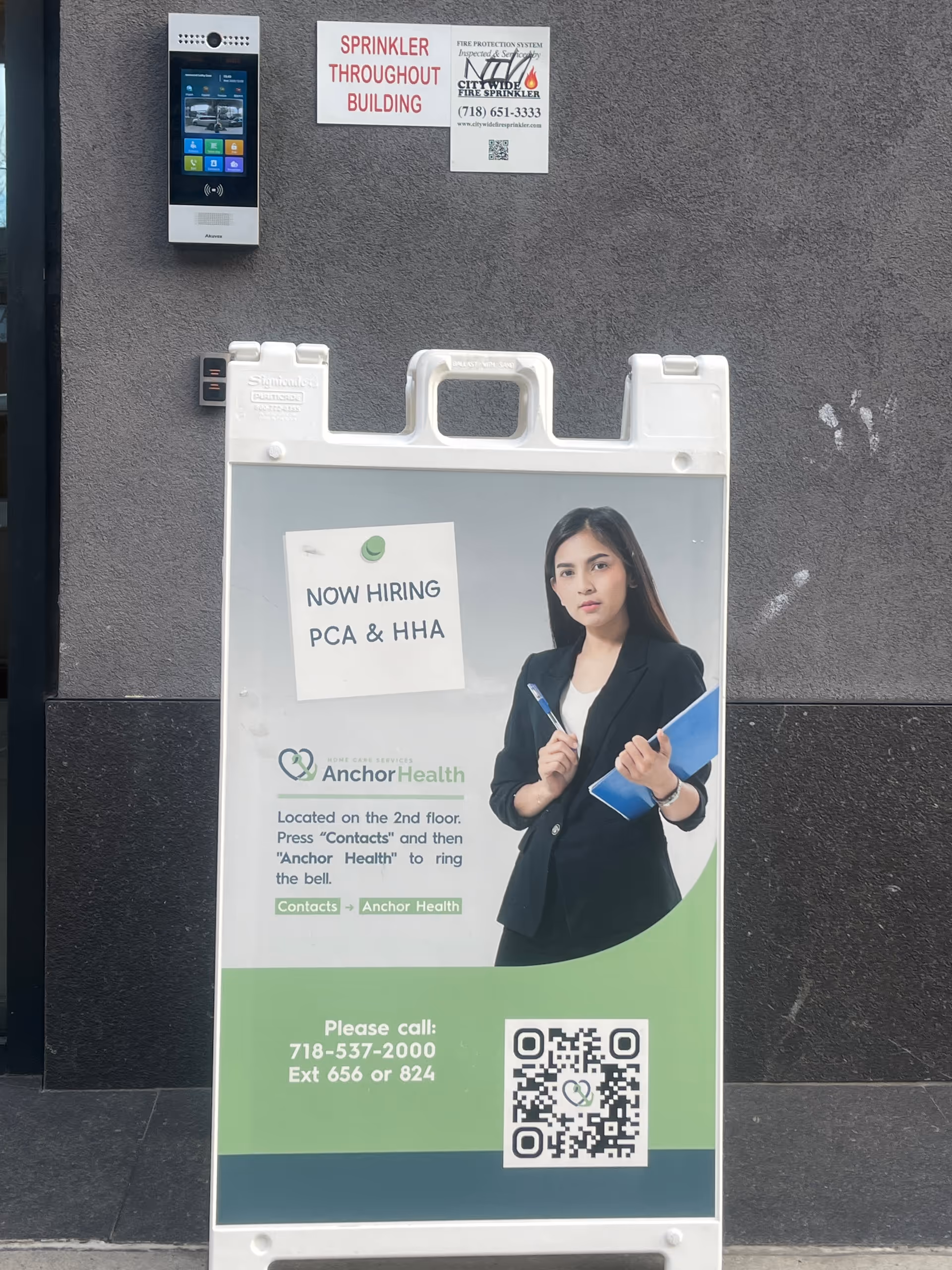Outdoor wall featuring Anchor Health ad sign, QR code, and a hiring photo beside an intercom.