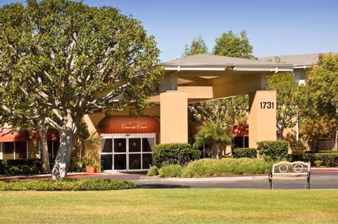 Emerald Court senior living facility entrance with red awning, beige pillars, trees, bench, and lawn.