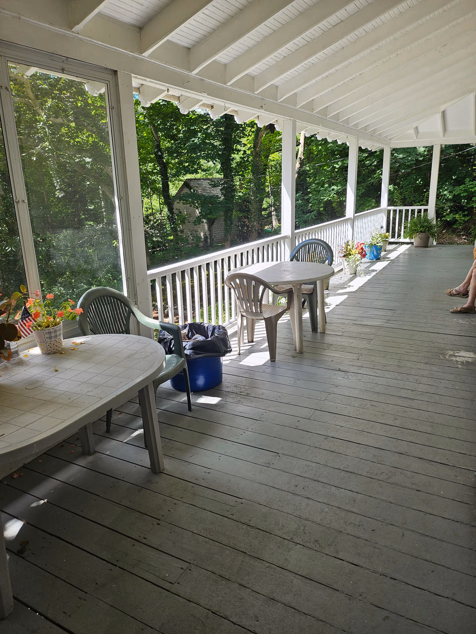 Covered wooden porch with tables, chairs, potted plants, and leafy trees surrounding.