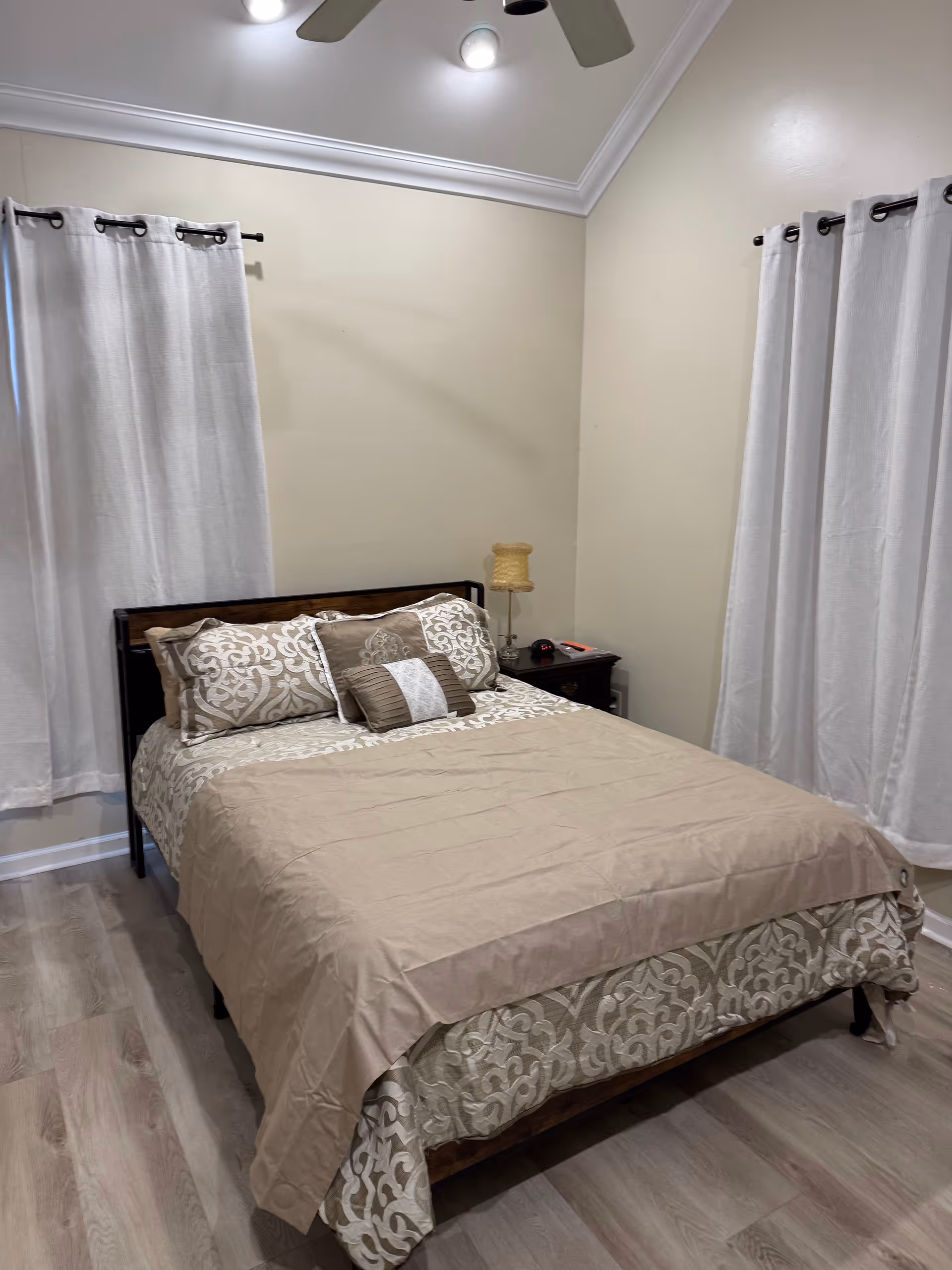 A neatly made bedroom with beige bedding, pillows, nightstand, and white curtains.