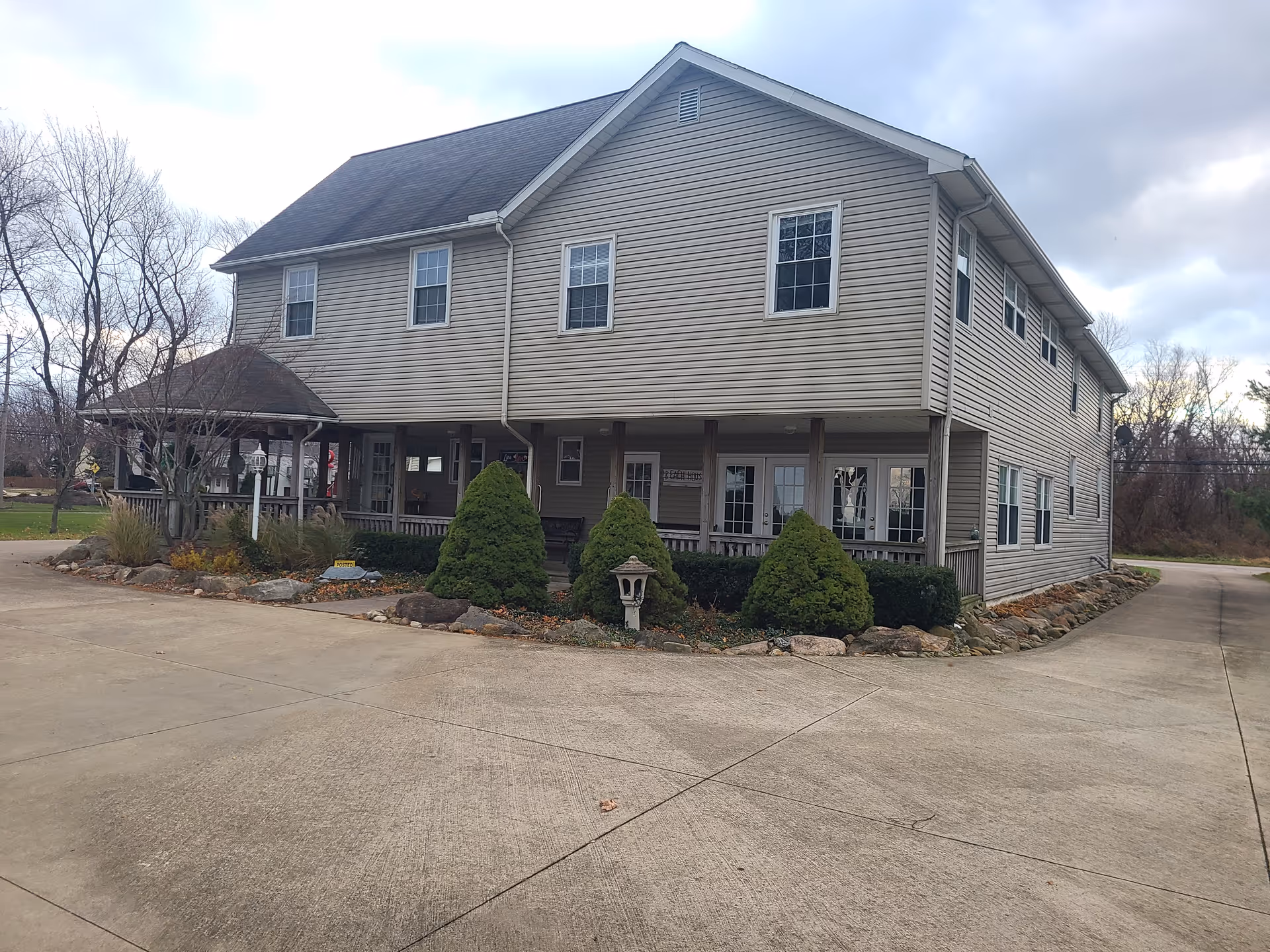 Two-story beige senior living facility with a wraparound porch and landscaped front yard.