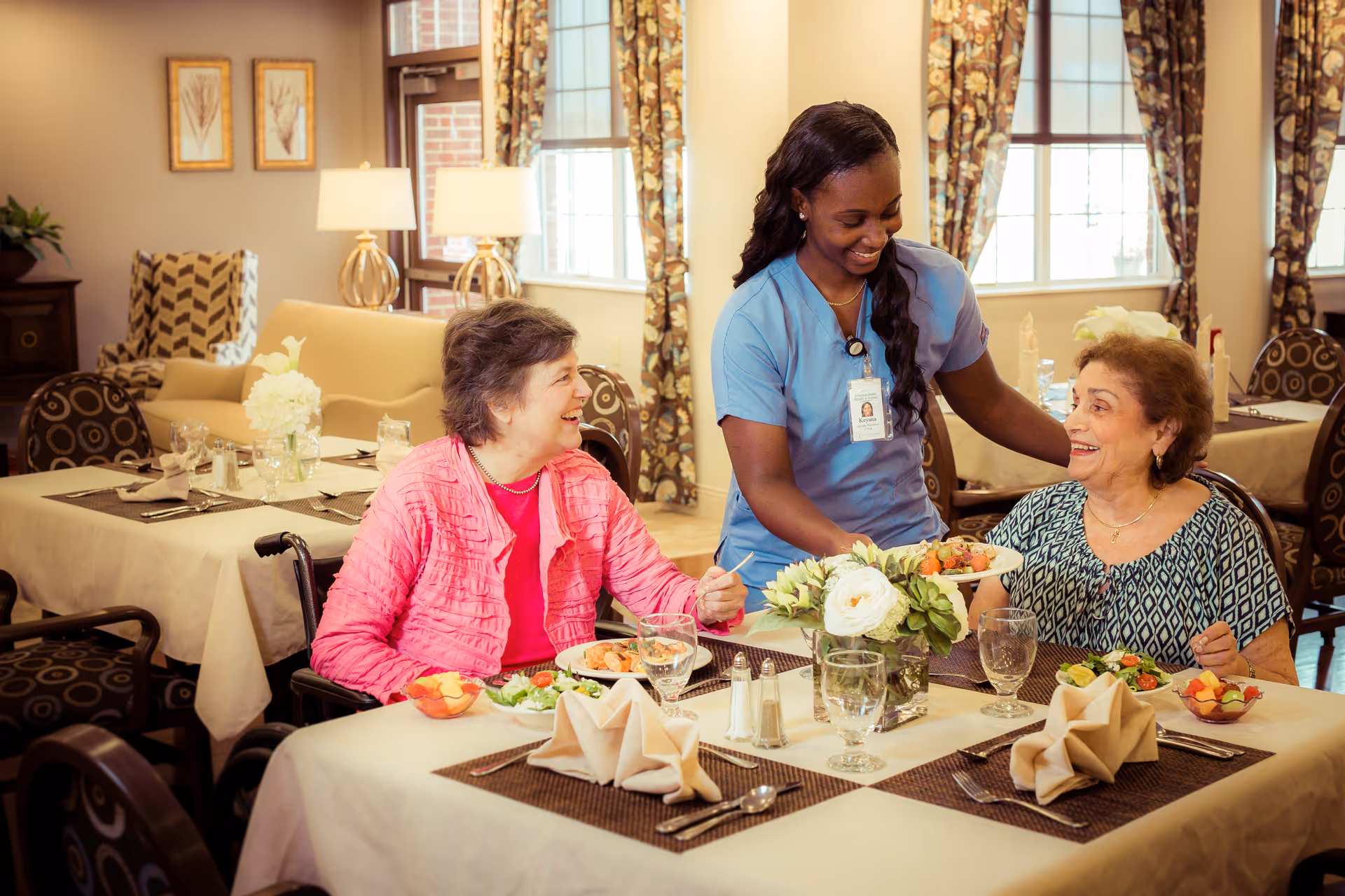 A smiling caregiver in blue scrubs serves a plate of food to an elderly woman seated at a dining table, while another elderly woman in a wheelchair sits nearby, all in a warmly lit dining room with tables set with flowers, glasses, and napkins.