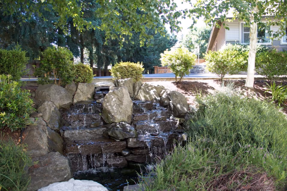 Tiered rock waterfall in a landscaped outdoor area with shrubs, trees, and part of a building visible in the background.