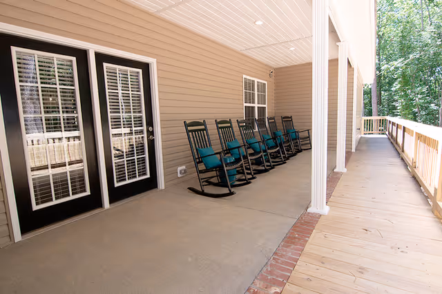 Covered porch with a row of black rocking chairs, double glass doors, and a wooden ramp overlooking trees.