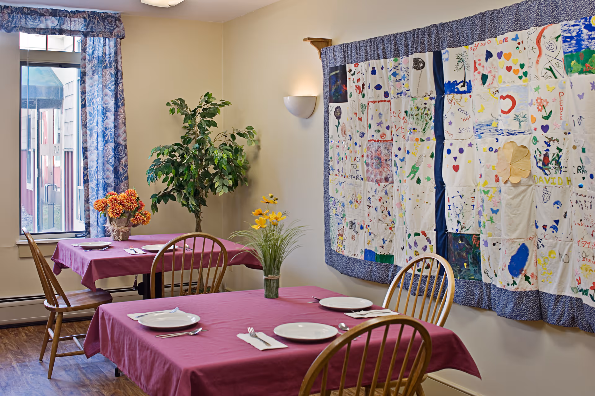 A dining room with two tables covered in purple tablecloths, each set with white plates, silverware, and napkins. Wooden chairs surround the tables. A large colorful quilt with various drawings and patterns hangs on the wall. A window with blue patterned curtains lets in natural light, and there are two vases with flowers and a potted plant in the corner.