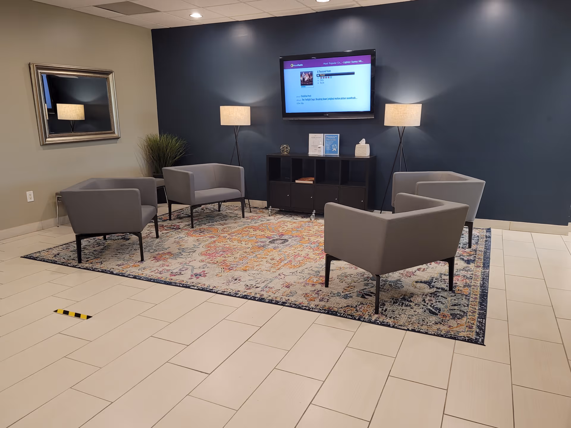 A modern waiting lounge with four gray chairs, a rug, and a wall-mounted TV.
