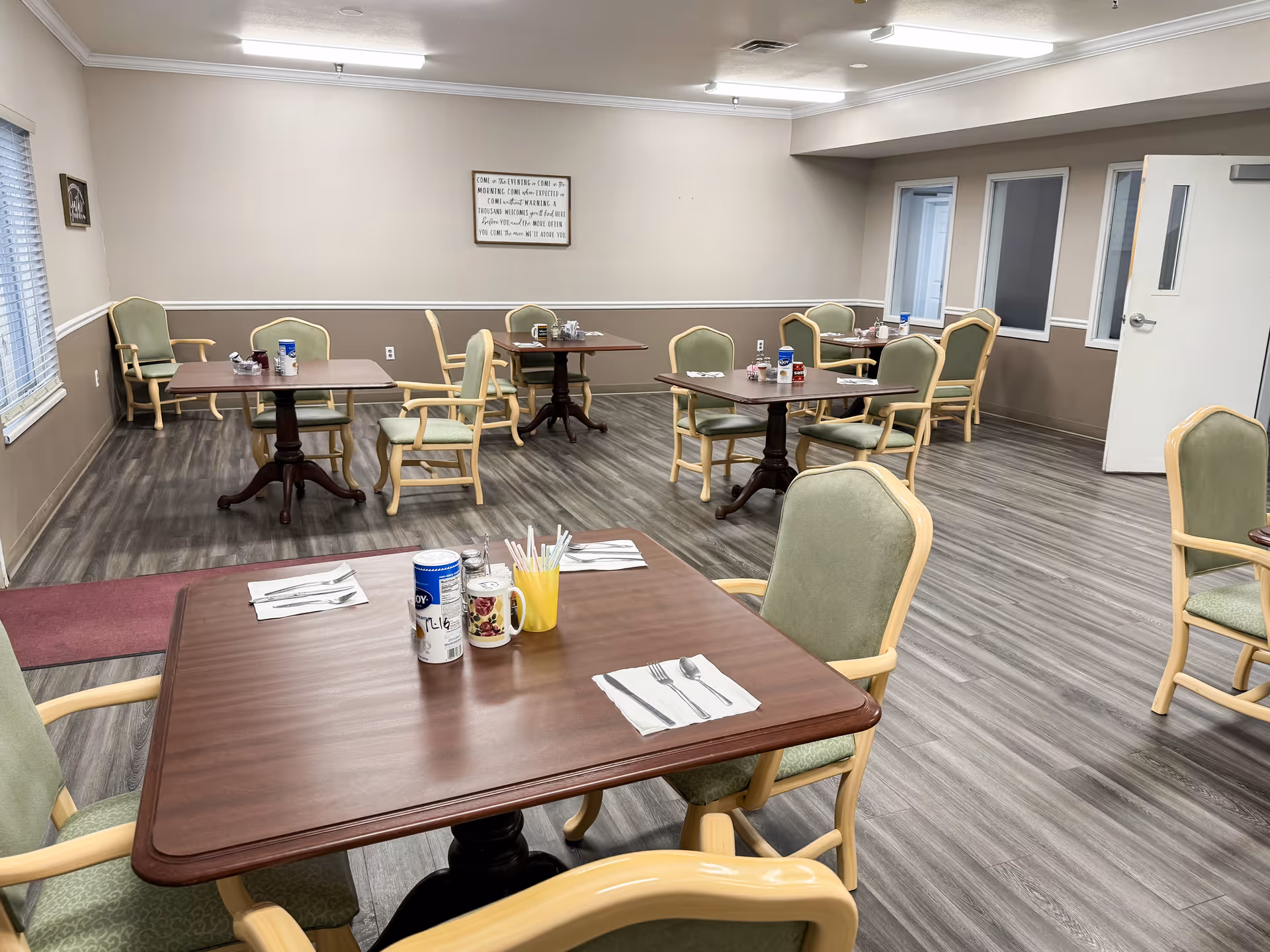 Senior living dining room featuring several tables, green upholstered chairs, and utensils.