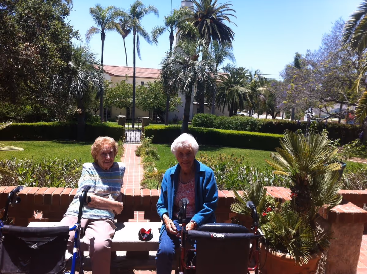 Two elderly women sitting on a bench in a garden area with palm trees and greenery around them. Both women have walkers in front of them. The background shows a building with a red-tiled roof and a clear blue sky.