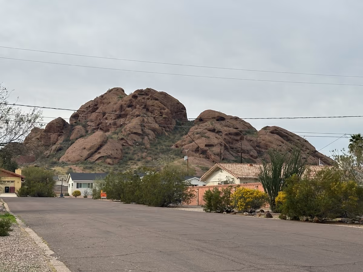 A quiet residential street with houses and desert vegetation in the foreground, and a large rocky hill or mountain in the background under a cloudy sky.