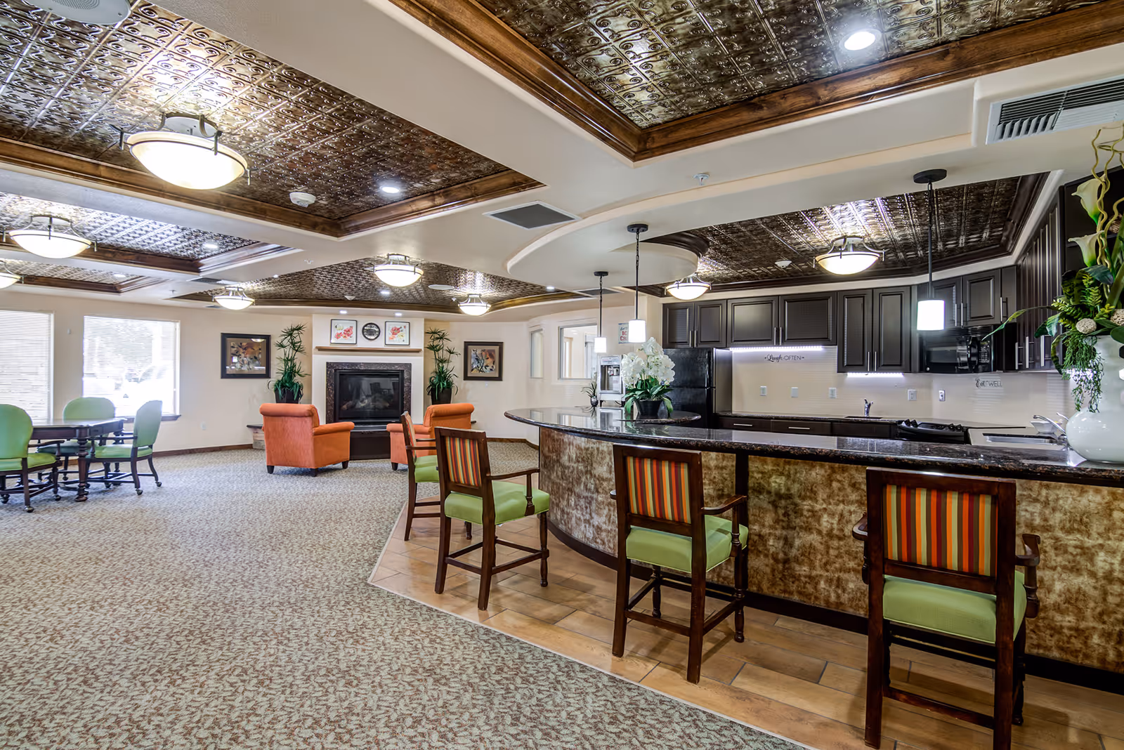 Bright senior living common area with a curved bar and high-backed chairs, seating area with armchairs and a fireplace, and decorative tin ceilings.