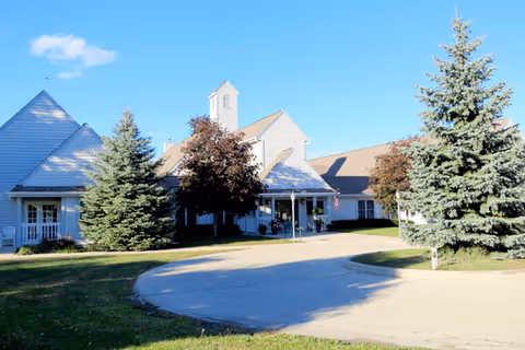 Front exterior of Matthews Of Horicon with a circular driveway, entrance canopy, evergreen trees and an American flag under a clear blue sky.