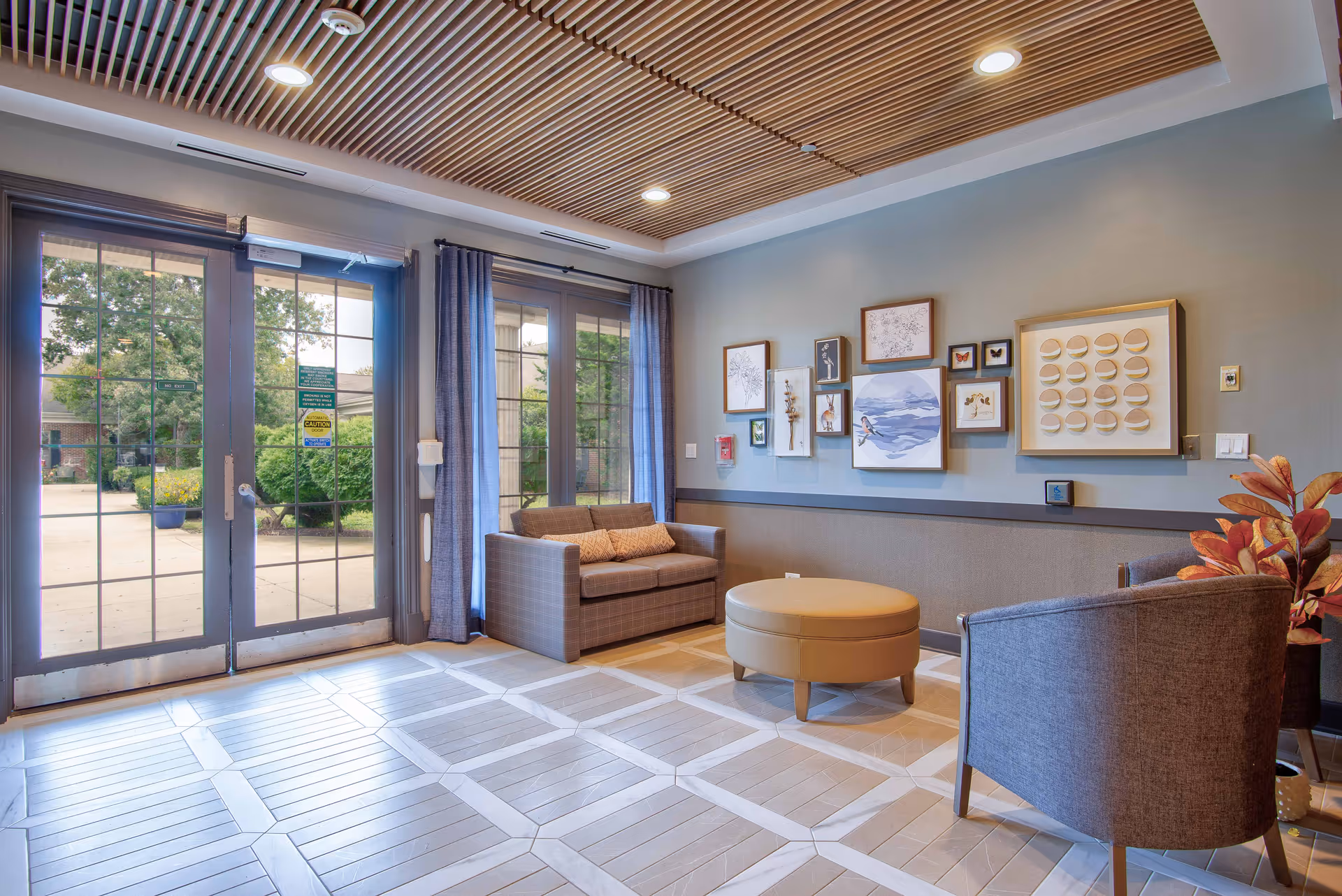 Lobby seating area with couches, an armchair and round ottoman, framed wall art and large glass doors looking out to a courtyard.