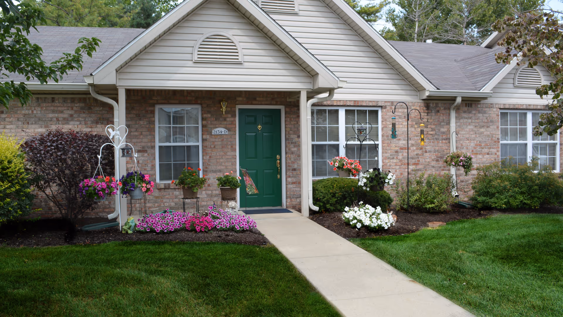 Exterior view of a single-story brick residential unit with a green front door, two windows with white blinds, and a concrete walkway leading to the entrance. The front yard is landscaped with green grass, various flowering plants, hanging flower baskets, and shrubs.
