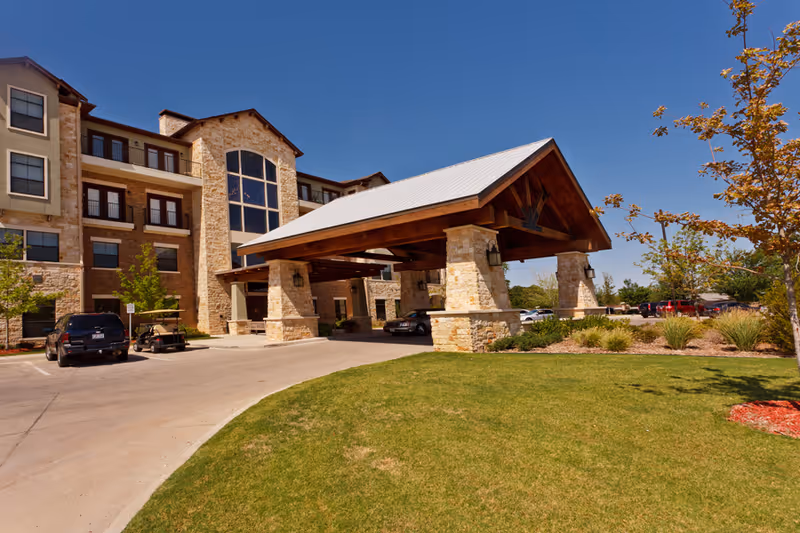 Exterior view of a multi-story senior living facility with stone and brick facade, large covered entrance with wooden beams, parked cars, and well-maintained landscaping under a clear blue sky.