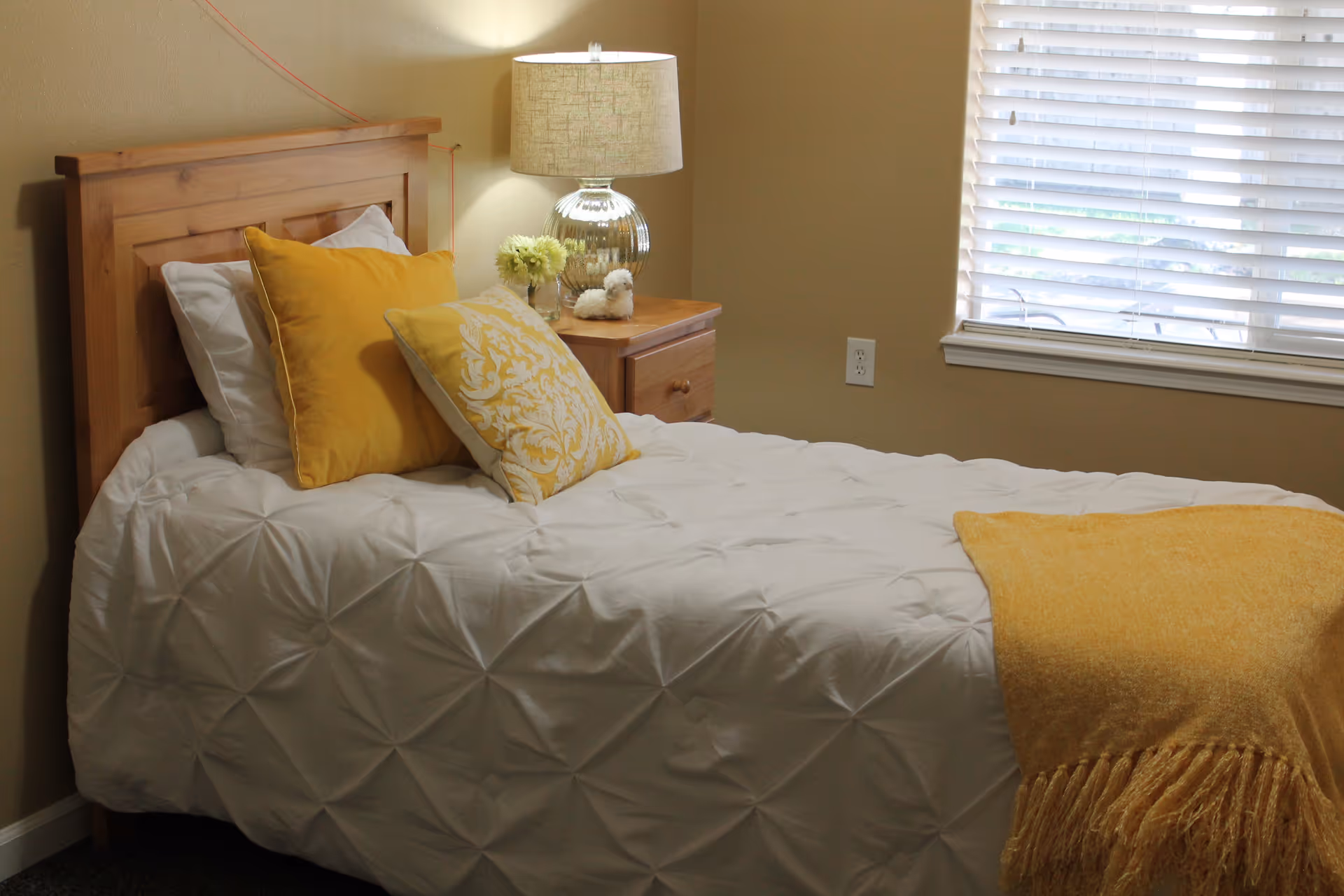 A neatly made bedroom with a wooden bed, yellow pillows, and a nightstand by a window.