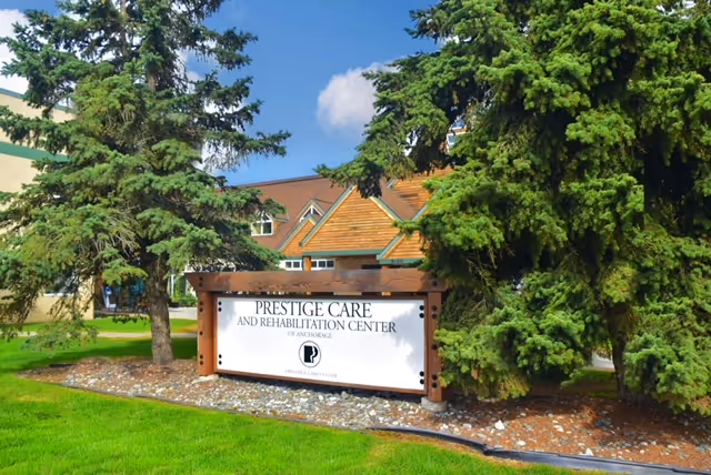 Outdoor view of the Prestige Care and Rehabilitation Center sign surrounded by green grass, trees, and a building with a brown roof in the background under a blue sky with some clouds.