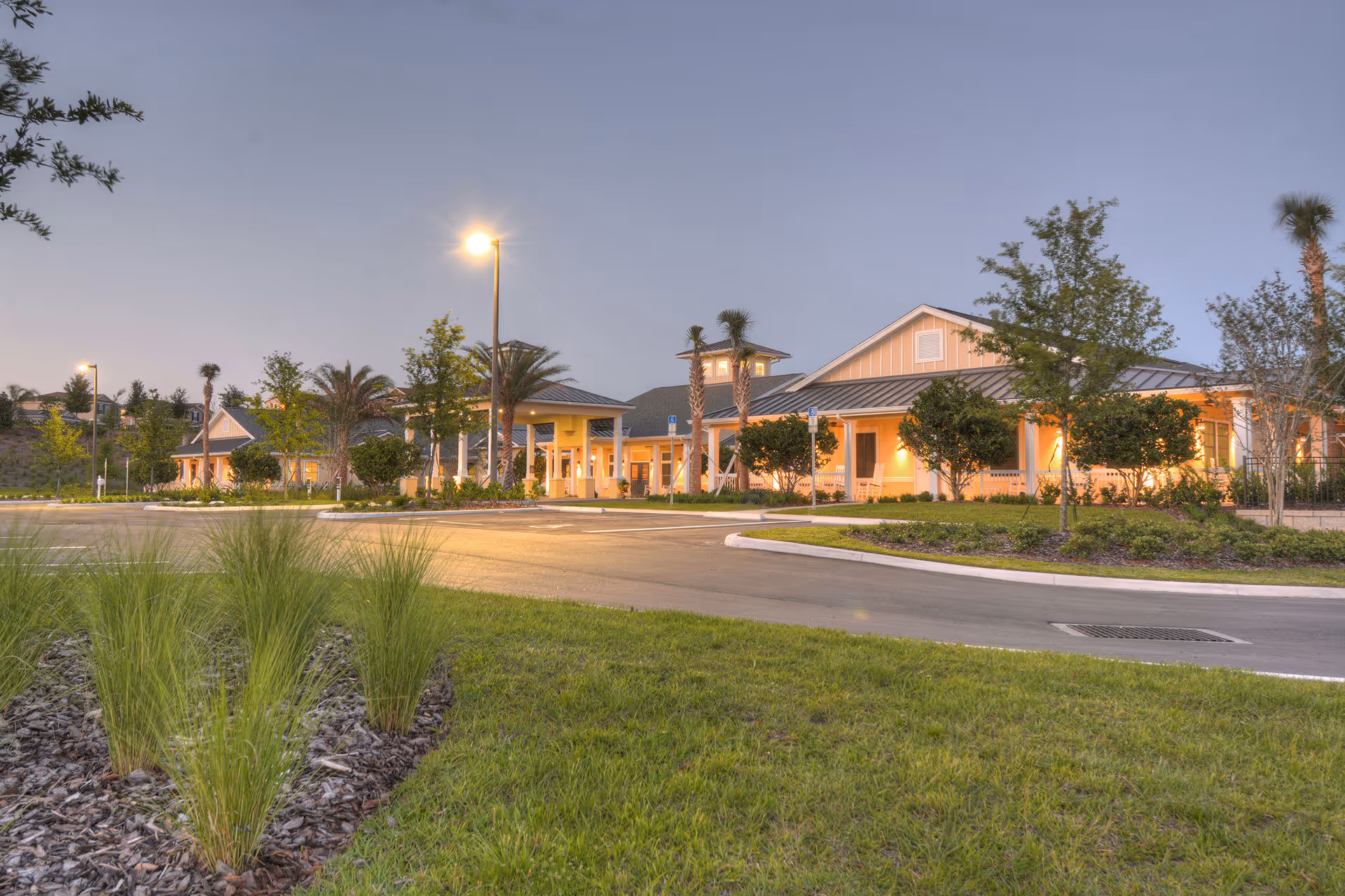 Exterior view of Benton House of Clermont at dusk, showing a well-lit building with a covered entrance, surrounded by landscaped greenery, palm trees, and a parking lot with street lamps.