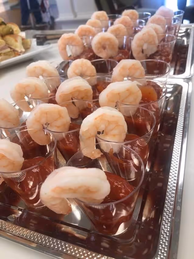 Individual shrimp cocktail servings in clear plastic cups with cocktail sauce arranged on a metal serving tray.