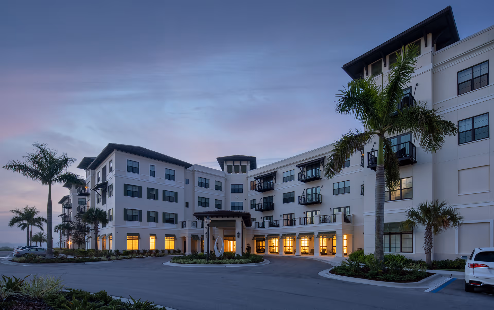 Exterior view of a modern senior living facility building at dusk with palm trees and a circular driveway. The building has multiple floors with balconies and large windows illuminated from inside.