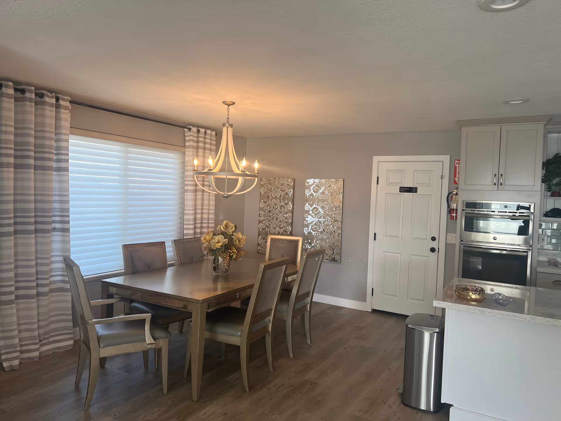 A dining area with a wooden table and six chairs, a chandelier hanging above the table, a vase with flowers on the table, striped curtains covering a window, two decorative wall panels, and a kitchen area with built-in ovens and a countertop visible to the right.