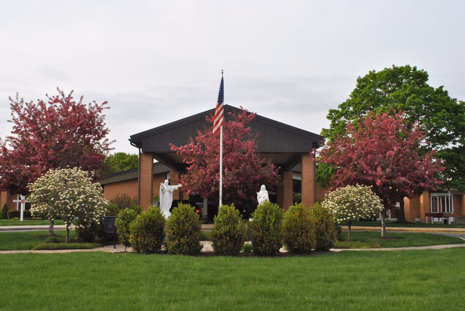 Front exterior view of St. Ann's Home featuring a brick building with a covered entrance, an American flag on a flagpole, two white statues, and landscaped greenery including flowering trees and bushes.