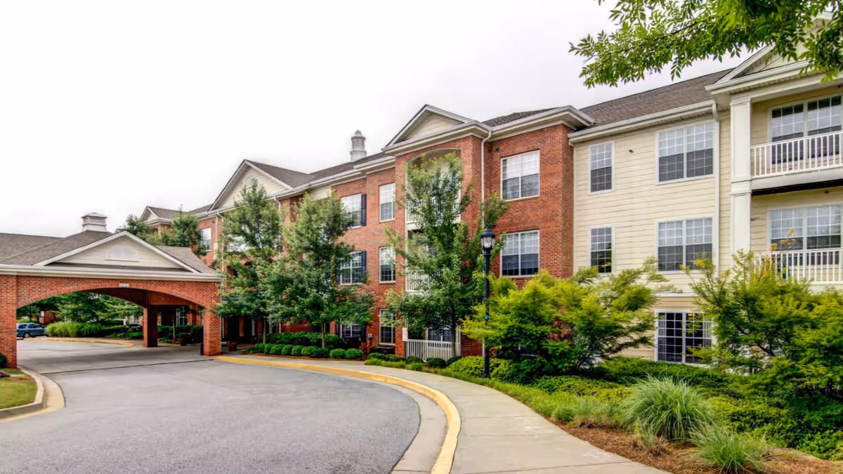 Exterior view of a senior living facility building with a covered driveway entrance, multiple windows, and a mix of red brick and beige siding. The area is landscaped with green bushes and trees along a curved sidewalk and driveway.