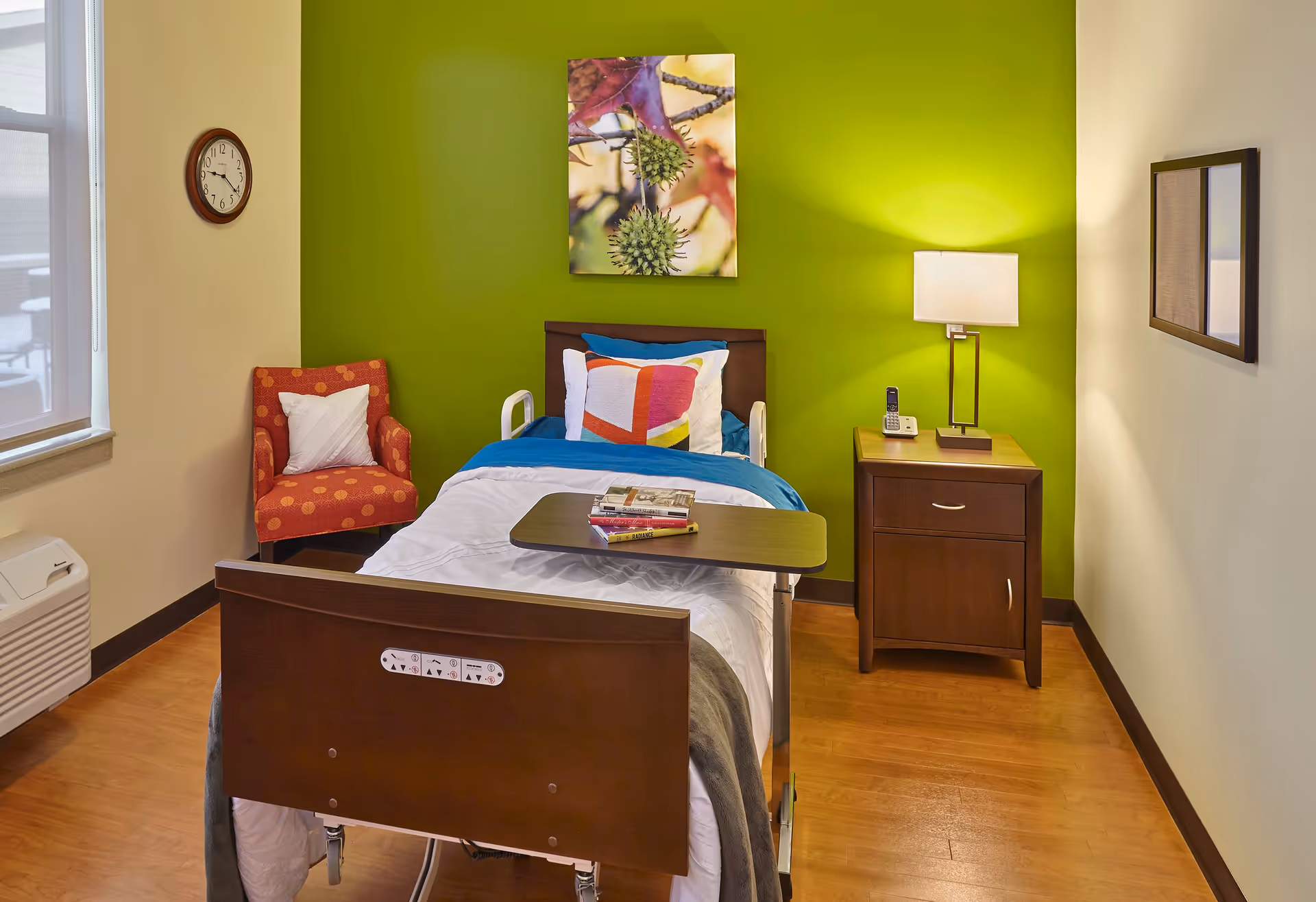 A small, cozy bedroom with a single hospital-style bed featuring white and blue bedding and colorful pillows. There is a wooden nightstand with a lamp and a cordless phone on it. A red armchair with a white pillow is placed near a window with a clock on the wall above it. The back wall is painted bright green and has a nature-themed artwork hanging on it. The floor is wooden.