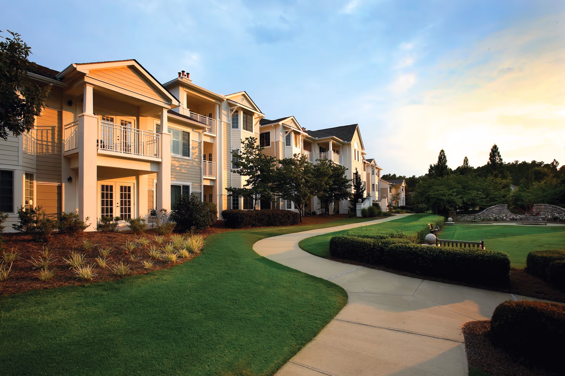 Exterior view of a senior living facility building with multiple balconies and large windows, surrounded by well-maintained green lawns, shrubs, and a paved walkway under a partly cloudy sky at sunset.