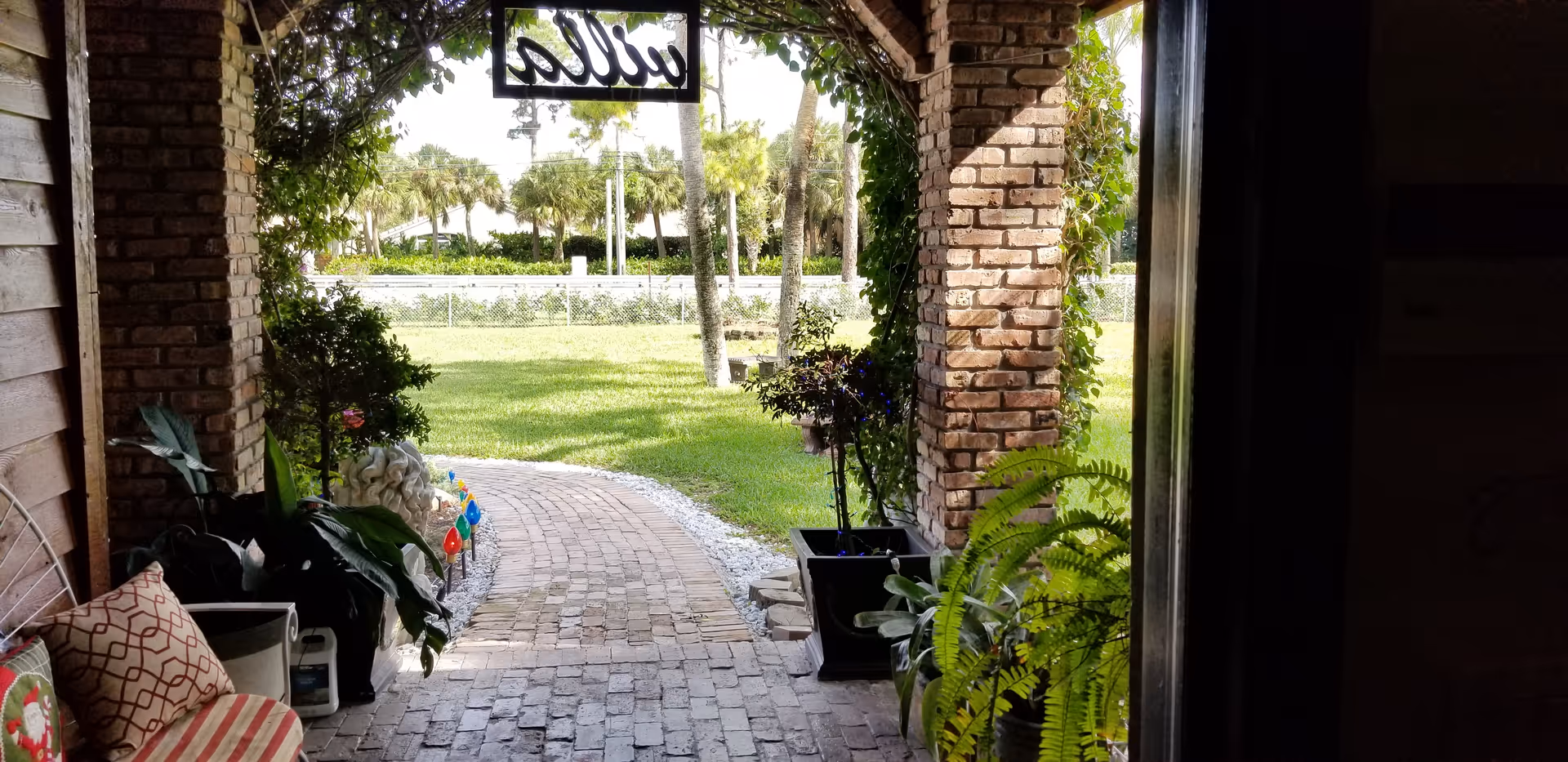 Brick porch with arches, hanging sign, potted plants, and a brick pathway to a green lawn.