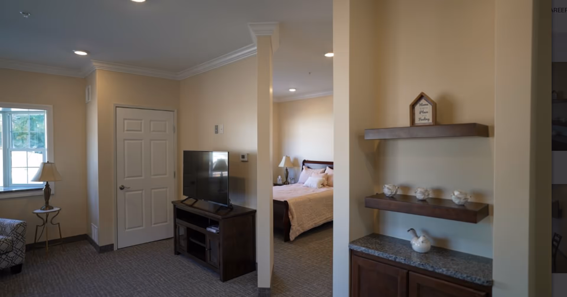 Interior view of a senior living facility room with a small living area featuring a patterned armchair, a side table with a lamp, a TV on a wooden stand, and a doorway leading to a bedroom with a bed, nightstand, and lamp. There are two wooden shelves on the right wall with decorative items and a small sign.