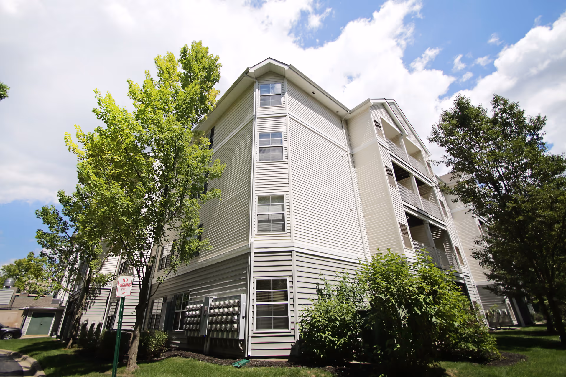Exterior corner view of a multi-story light-gray apartment building with balconies, trees, and a partly cloudy sky.