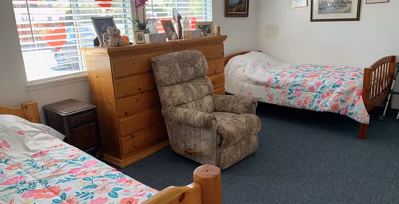 Shared bedroom with two single beds in floral bedding, a patterned recliner and a wooden dresser beneath a window.