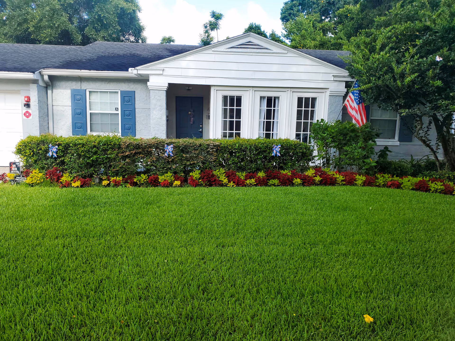 A single-story gray building with white trim, manicured lawn, hedges, flowers, and an American flag.