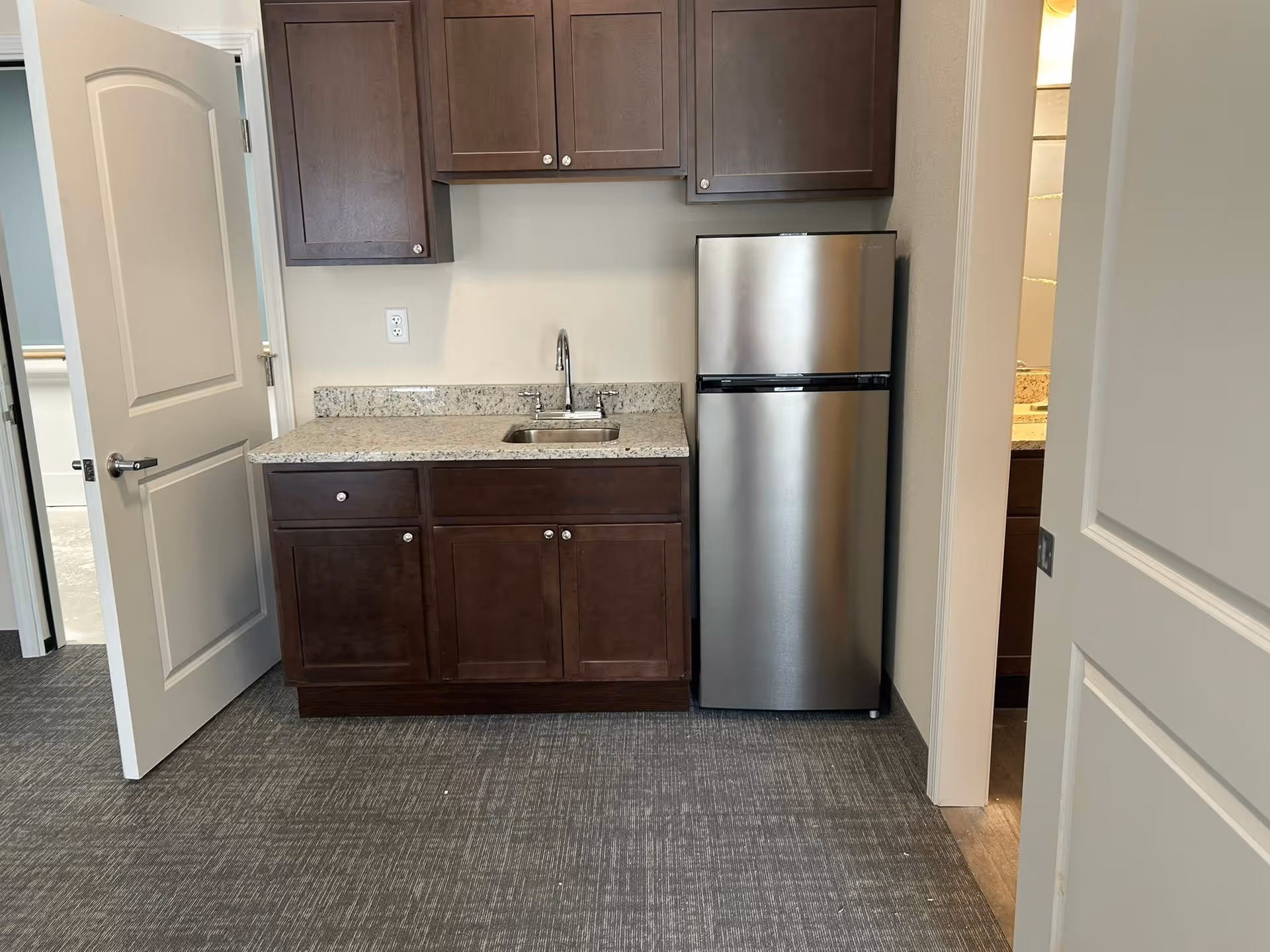 Small kitchenette with dark cabinets, granite countertop, sink, and stainless steel refrigerator.